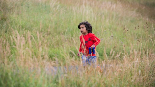 A small child wearing blue shorts and a red jumper, carrying a pair of binoculars, walks through tall, meadow grasses on a paved path.